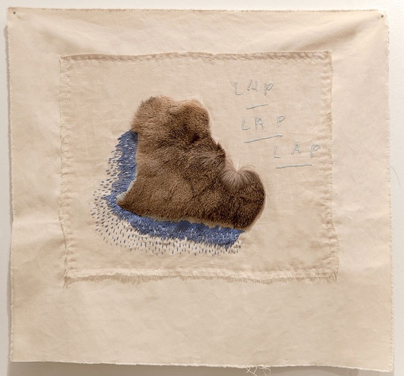 A square of linen with a piece of brown fur edged in blue embroidery, and faint stitched words above it.