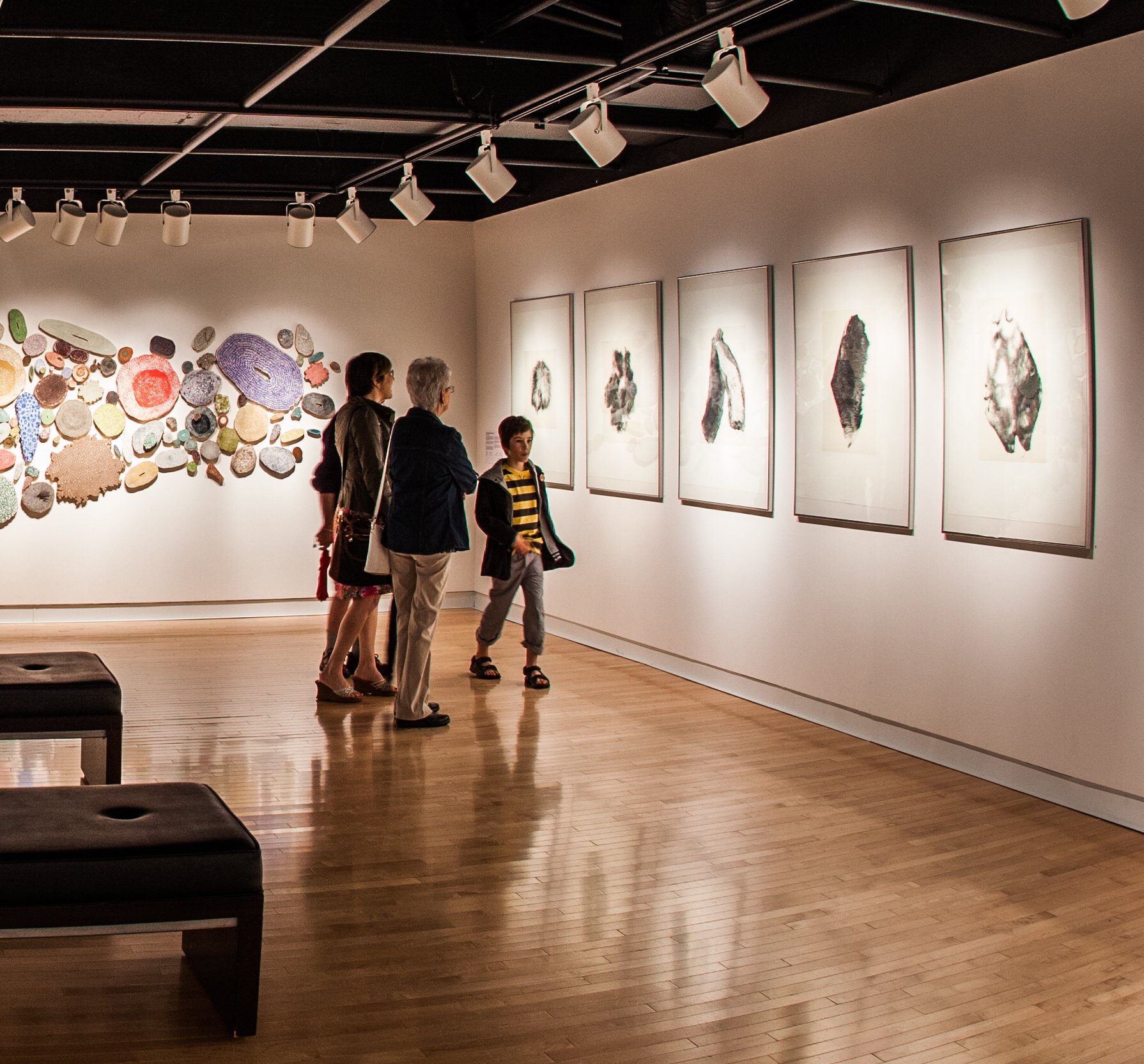 Installation view: five framed photopolymer etchings of fur remnants in a row on a lit gallery wall, with visitors looking at them.