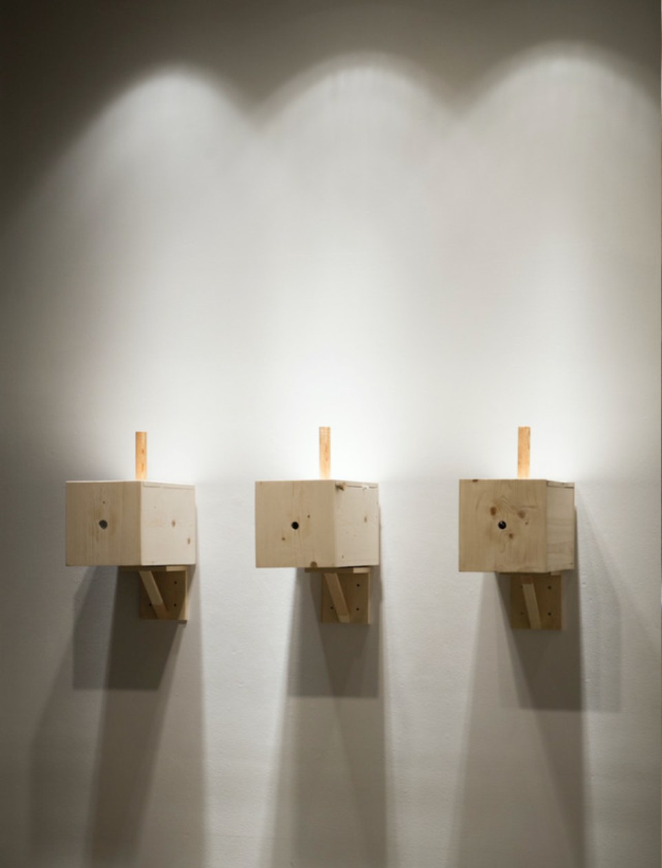 Three pale wooden boxes mounted on a white wall, each with a peep hole and a short wooden crank, lit from above.