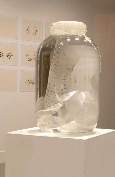 A glass jar on a plinth holding strips of gampi paper with handwritten text, with framed drawings of hands on the wall behind.