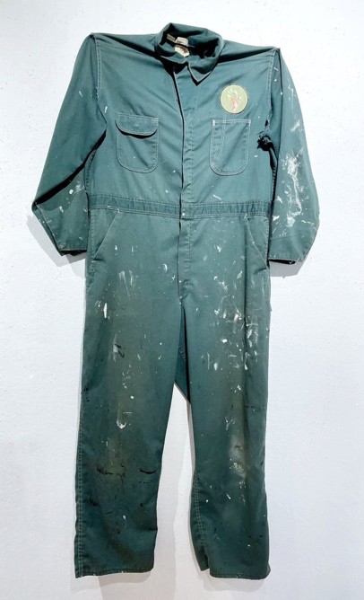 Green coveralls hanging on a wall, with a small embroidered patch on the chest.