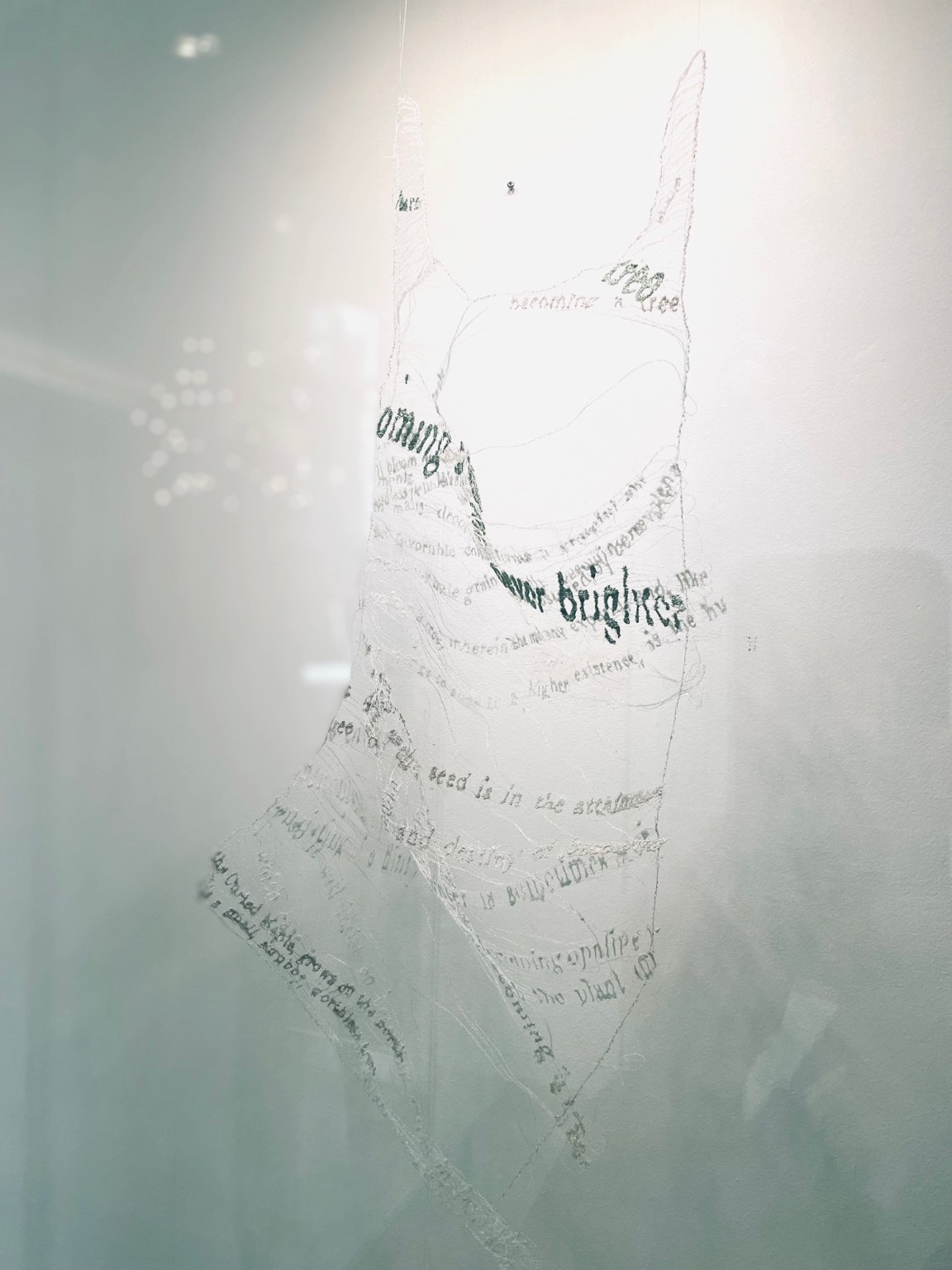 Machine-stitched lace hanging against a pale wall: a tall, narrow tree form made of stitched lines of text.