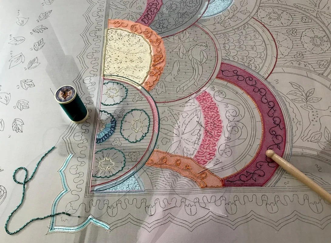 A Lunéville embroidery sampler in progress on the frame: outlined motifs, with areas filled in vermicelli, chain stitch and sequins; a spool of green thread and a tambour hook rest on the cloth.
