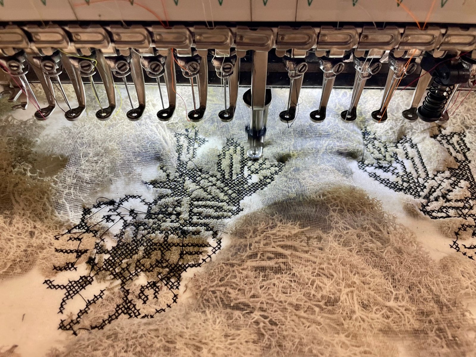 Black cross-stitch running on a loose gauze under the needles of a multi-needle embroidery machine, with earlier stitched passages around it.