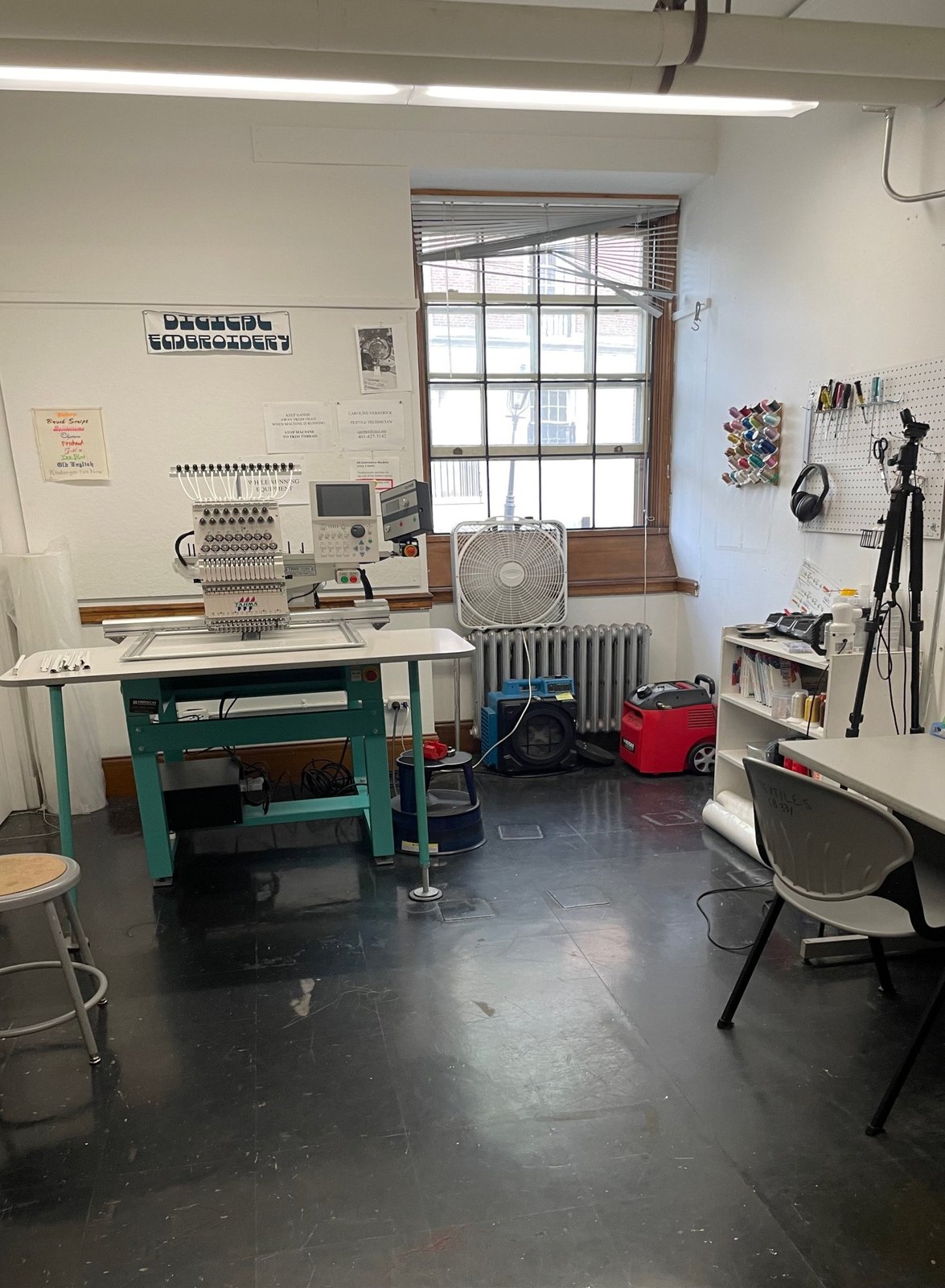 A small studio with a single-head Tajima embroidery machine on a green stand under a window, with a pegboard of tools and a workbench.